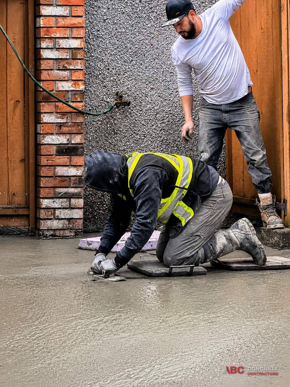 Two crew members hand-trowelling a fresh concrete slab beside an exposed aggregate wall